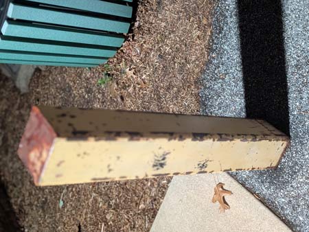 A rectangular yellow parking post casting a long shadow, a portion of a green paneled trash receptacle, and a portion of sidewalk alongside a black parking lot surface turned -90 degrees to create an abstract effect