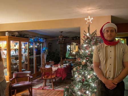 Will, an adult male, wearing a long flowing red stacking cap with white fur trim and a white pom at the end of the cap, standing in front of the right half of a decorated Christmas tree