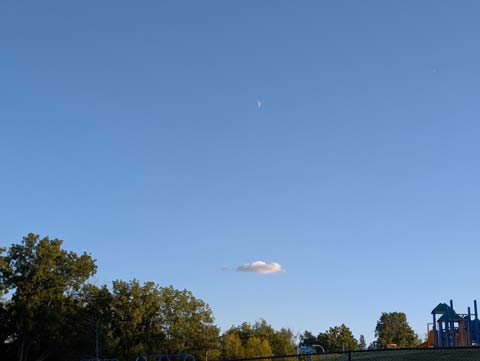 A lone cloud in a bright blue sky, with a crescent moon hovering high above the cloud 