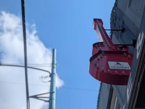 A red sign in the shape of a Gibson guitar, outlined on the inner edge with white lights, not turned on yet since the photo was taken in the daytime.