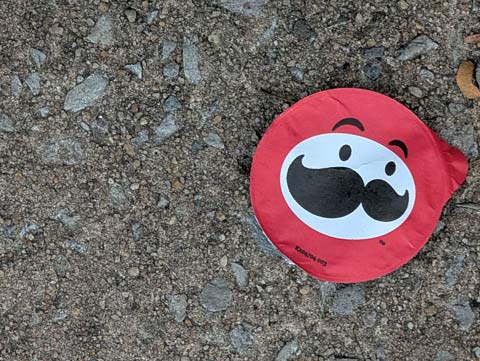 A red paper lid liner of a Pringles potato chip canister, with the face of Julius Pringle, the Pringles mascot character, laying discarded on a sidewalk