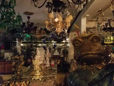 View through glass front door of curio shop, with human size frog statue standing, appearing to be ready to greet customers