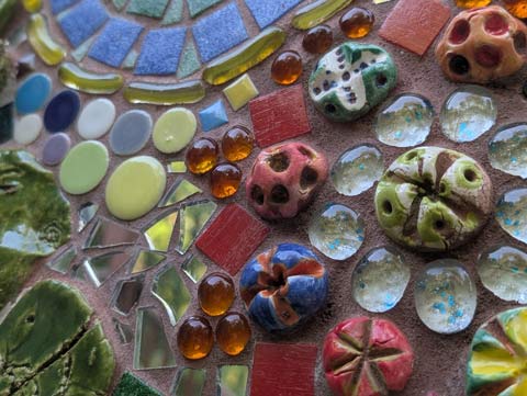 Close-up of a wall mosaic showing a variety of tile pieces of different shapes and colors, along with a variety of glass beads and ceramic flower heads