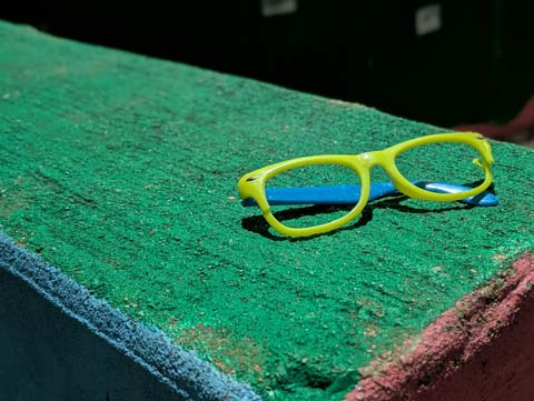 A pair of yellow and blue, lenseless, plastic eyeglasses, sitting on a cement wall painted green on top, blue one onse side, and red on the other.