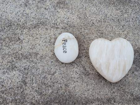 A white heart shaped stone, sitting beside another oval shaped stone with the word "PEACE" engraved on it, on the top of a grey granite tombstone