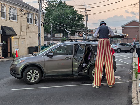 Rear view of a man standing on stilts wearing red and white striped pants next to a park car with a door open