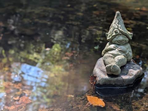 A small cement figure of a napping gnome, laying atop a rock, in the middle of a stream