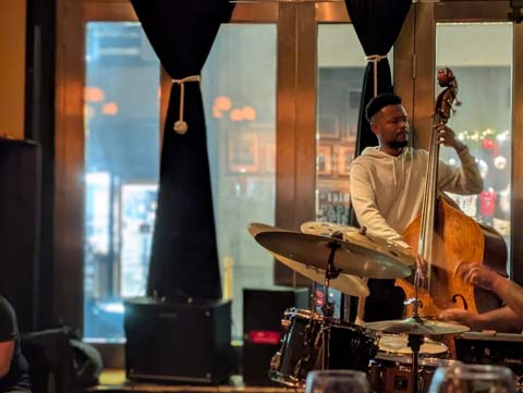 A man playing an upright bass, with a view of a city street through the floor to ceiling window behind him