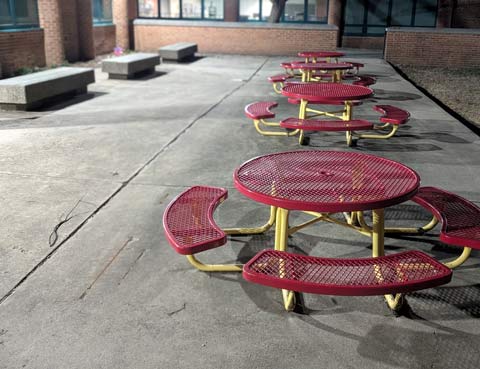 A line of four round metal tables each with four attached curved benches. The benches and table tops are red, the pipes that hold the table and benches together are yellow